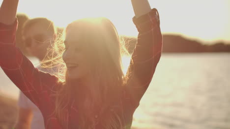 A-female-student-is-dancing-on-the-open-air-party-with-her-friends-on-the-beach.-Her-long-blonde-hair-is-flying-on-the-wind.-She-smiles-and-enjoys-a-summertime-on-the-river-coast-with-beer.