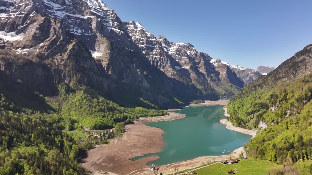 Aerial view of Klöntalersee in Glarus, Switzerland, surrounded by steep snow-capped peaks and lush green forests under a clear blue sky