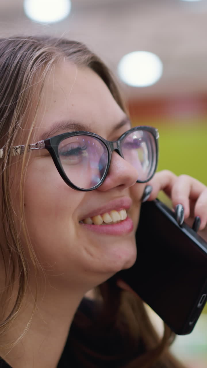 vista lateral de primer plano de una mujer alegre hablando por teléfono con camiseta negra y gafas, con fondo borroso con efecto de luz bokeh y estructuras modernas