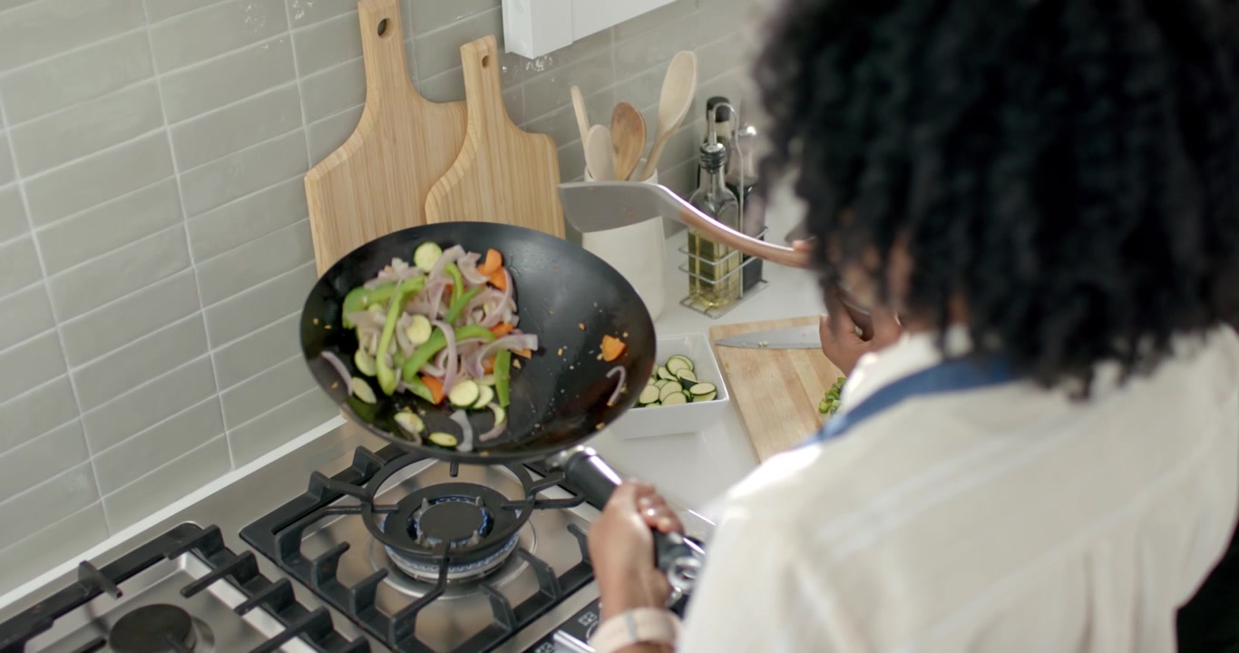 Couple cooking in kitchen with prepped veg, woman holding wok tossing stirfry as man seasoning pan