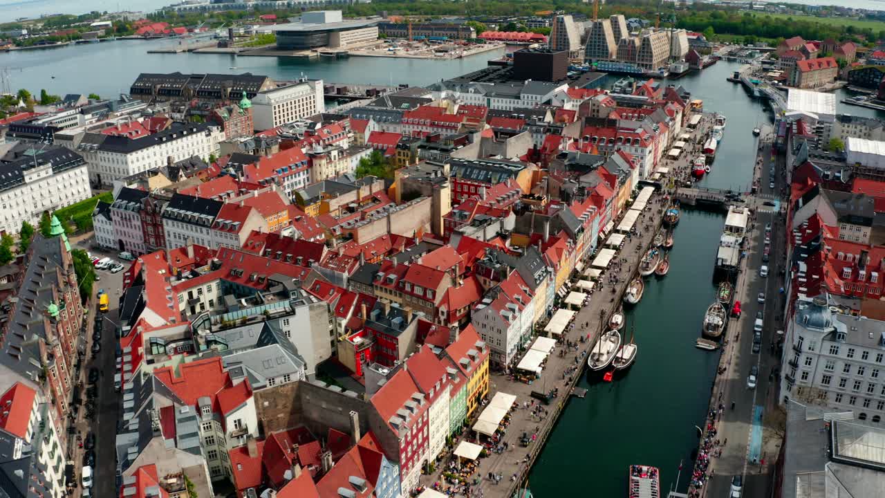 High view of the iconic Nyhavn street scene with brightly coloured townhouses, a canal with historic boats, and tourists enjoying the sunny day out on the waterfront.