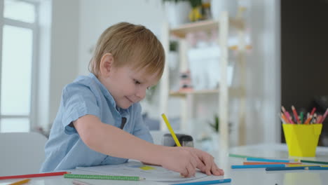 Lächelnder-Junge-Im-Blauen-Hemd-Zeichnet-Mit-Einem-Bleistift-Auf-Papier,-Während-Er-Am-Tisch-Im-Wohnzimmer-Sitzt