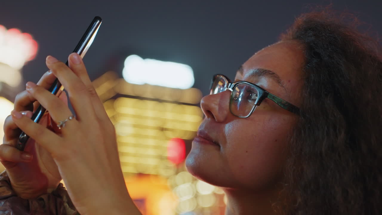 Black woman using smartphone at night with city bokeh and warm glow. Closeup portrait shows glasses and curly hair, hands holding device, smartwatch on wrist, focused expression scrolling messages