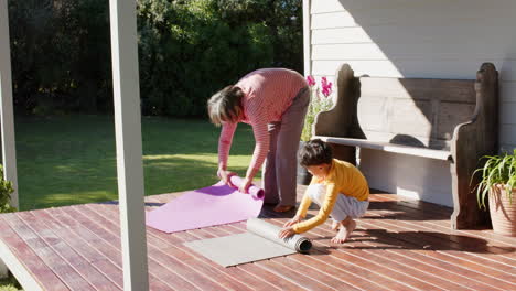 Abuela-Birracial-Mayor-Y-Nieto-Rodando-Esteras-De-Yoga-En-La-Terraza,-Cámara-Lenta