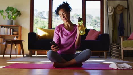 Mixed-race-woman-drinking-water-and-using-smartphone-while-sitting-on-yoga-mat-at-home