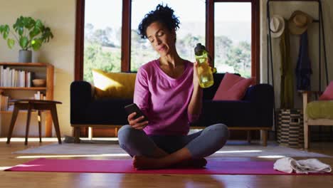 Mujer-De-Raza-Mixta-Bebiendo-Agua-Y-Usando-Un-Teléfono-Inteligente-Mientras-Está-Sentada-En-Una-Estera-De-Yoga-En-Casa