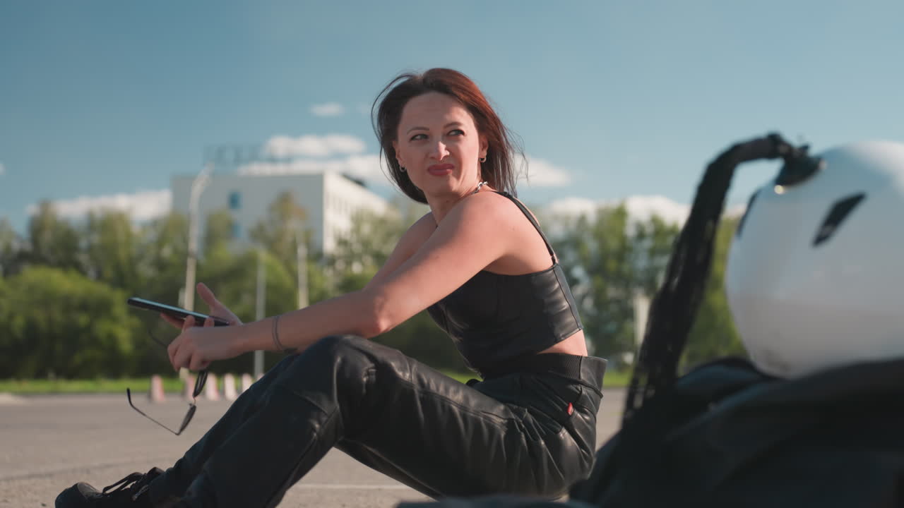 Close up of helmet beside woman seated on pavement flaunting hair back placing hand on chin while holding sunglass and typing on phone under bright daylight with blur urban background