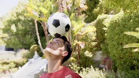 hombre biracial enfocado practicando fútbol de equilibrio en la cabeza en un jardín soleado, cámara lenta