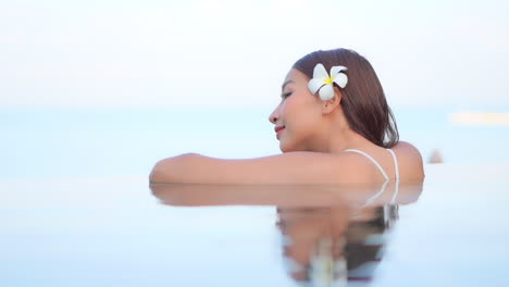 a close-up of an attractive woman with a flower behind her left ear leans along the edge of an infinity pool edge