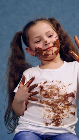 happy kid with dirty hands and face by chocolate. funny little girl has smeared face, hands and white t-shirt with chocolate in the studio. vertical video