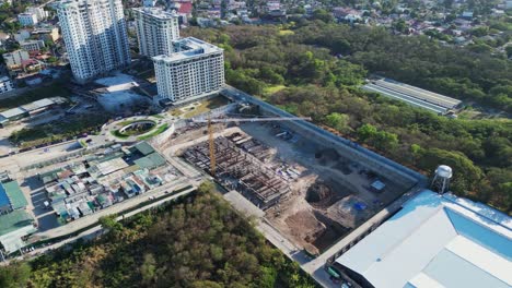 Overview-of-city-construction-area-with-large-crane-surrounded-by-buildings-and-trees-in-Alabang,-Muntinlupa-City,-Philippines--aerial-pullback-shot