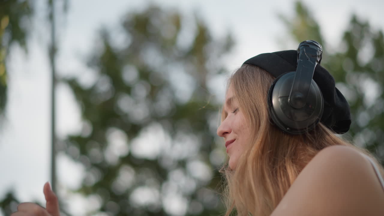 Giovane donna bianca che balla all'aperto con le cuffie, spensierata ballerina di strada che si muove a ritmo di musica in un parco urbano alberato, cuffie nere oversize, canotta bianca casual, lunghi capelli biondi, mani espressive