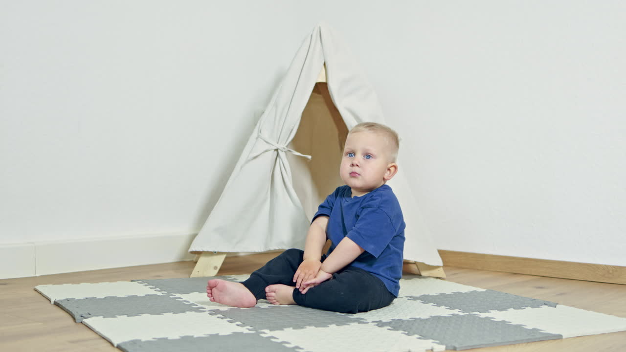 Niño pequeño alegre jugando en casa sobre una alfombra y sentado en un tipi. Una casita para un niño en un apartamento. Un tipi para un niño. Un niño en la sala de estar es travieso y se ríe.