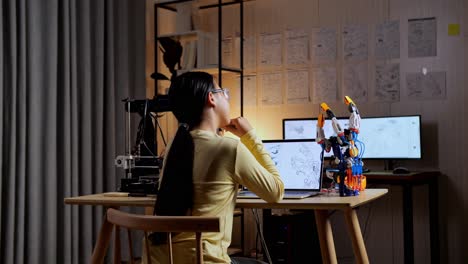 back view of teen asian girl with 3d printing thinking and raising index finger while designing a cyborg hand on a laptop at home