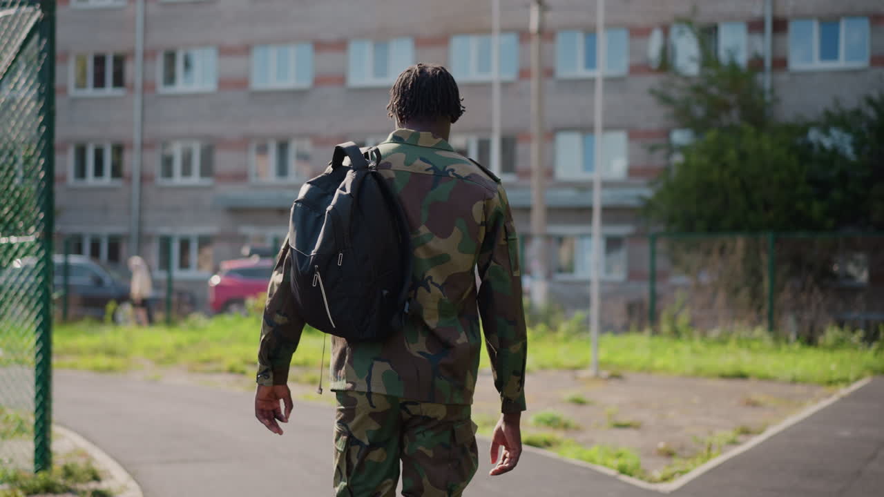 Soldier Leisurely Strolling Past Housing Units, Relaxed Soldier Ambles Along Fencelined Street Behind Apartments, Soldier Enjoys Calm Walk Along Fenced Pathway With Apartment Buildings In Distance