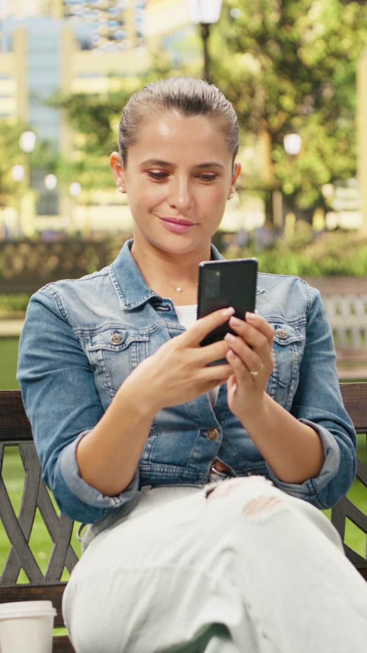 Video verticale: donna sorridente usa lo smartphone mentre si rilassa su una panca di legno.
