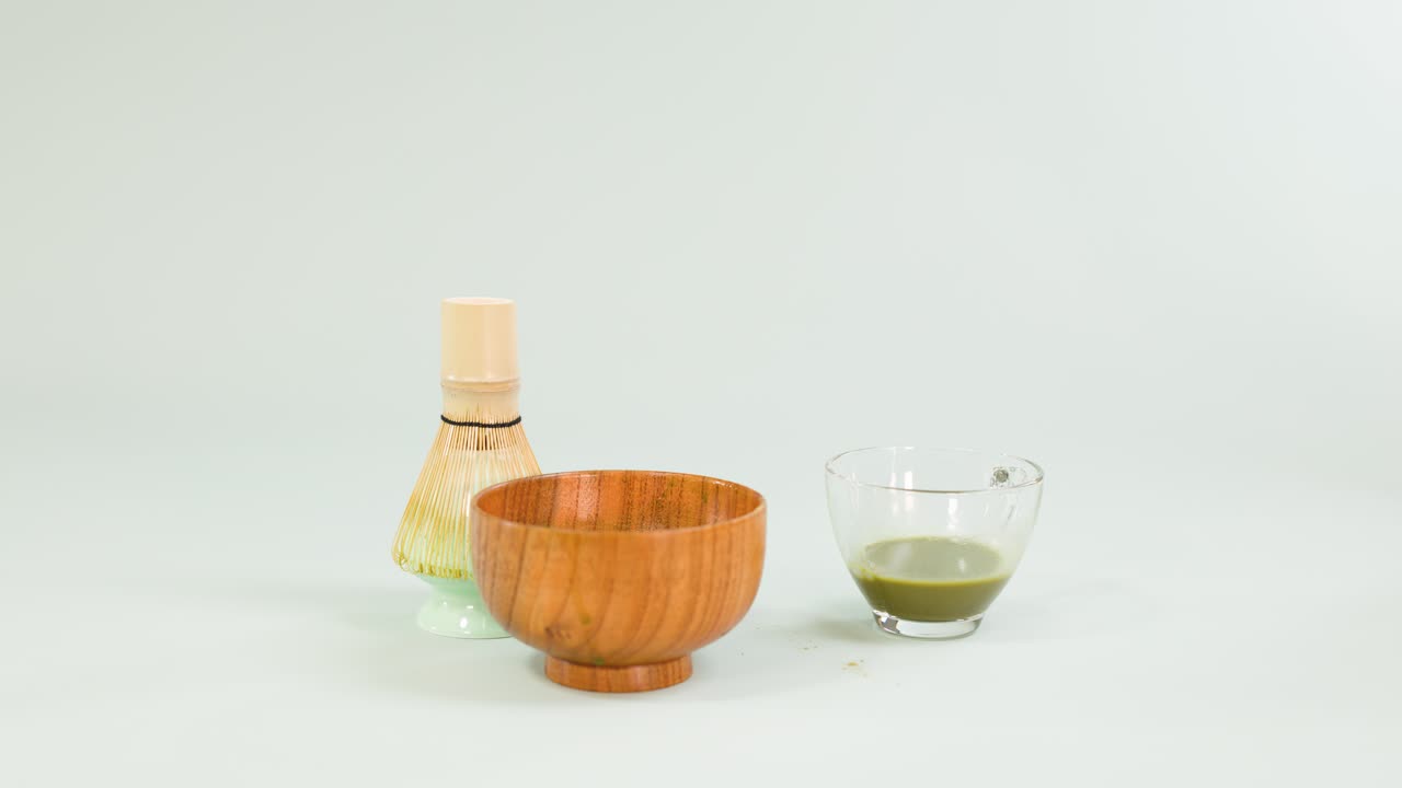 Hand pours water into matcha tea glass beside bamboo whisk, wooden bowl, minimal clean background