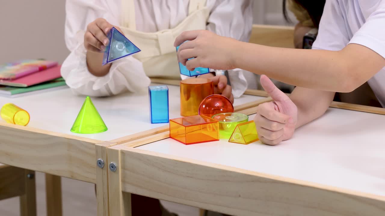 Adult and child stack transparent geometric blocks on desk in bright classroom, static framing