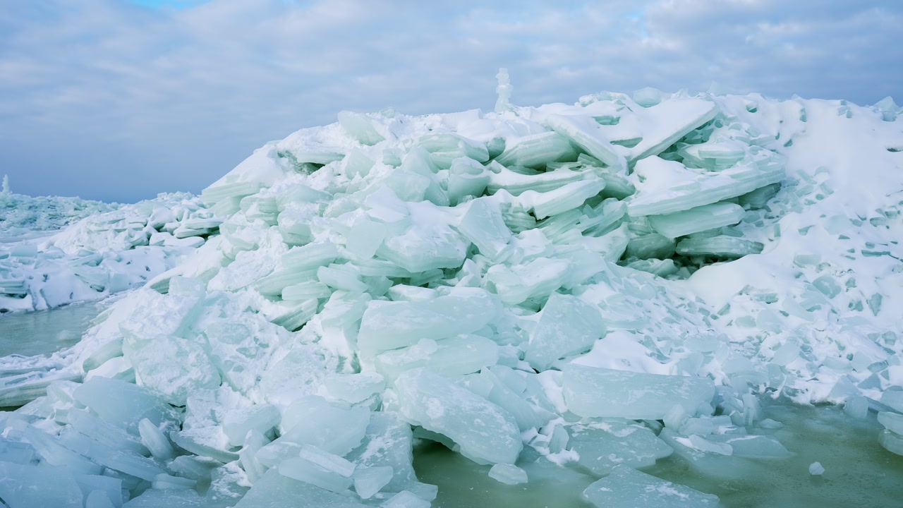 Close view of a massive sea-ice pressure ridge with blue ice slabs and slushy meltwater under a cloudy sky. Dramatic arctic texture showing fragile winter conditions.