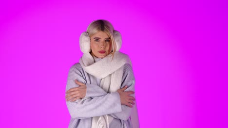 portrait of young blonde woman feeling cold, wearing scarf grey sweater and earmuffs, studio shot with pink background