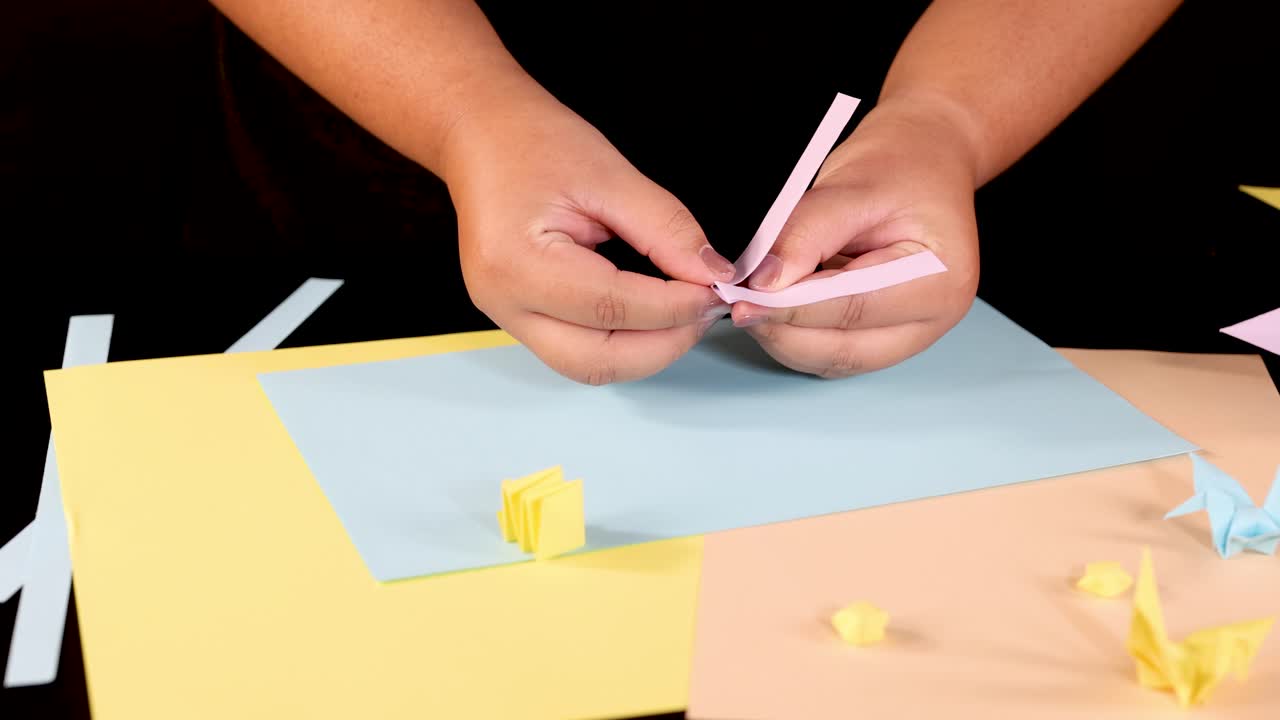 Person folds pastel paper strips into origami shapes on a well-lit, colorful workspace