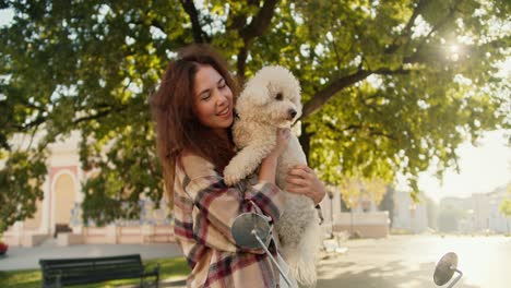 A-happy-brunette-girl-in-a-plaid-shirt-holds-a-curly-small-white-dog-in-her-arms,-smiles-and-looks-at-the-camera-in-a-summer-city-park-near-her-moped