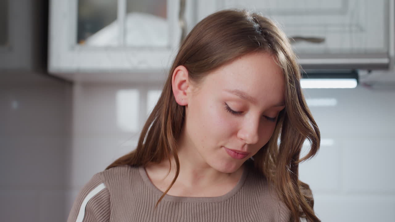Beautiful young woman with wavy brown hair and stylish top focuses on something while moving her head slightly, she enjoys a moment in a modern kitchen with soft lighting