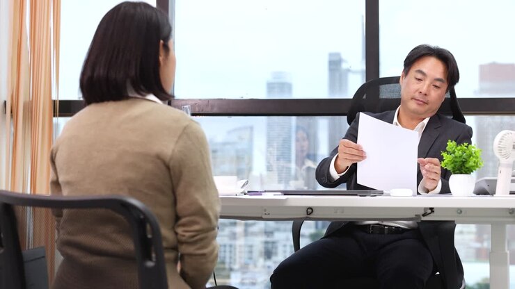 Professional Asian businessman reviews paperwork while interviewing a female candidate in a bright corporate office