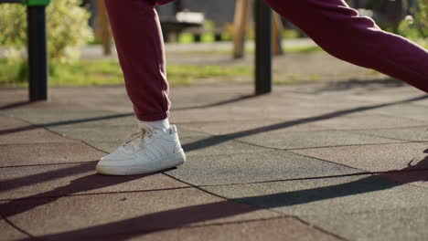 female athlete in hoodie and quilted vest grips iron bar while lunging forward on modern urban park tiles under sunny sky casting long shadows with relief expression on her face