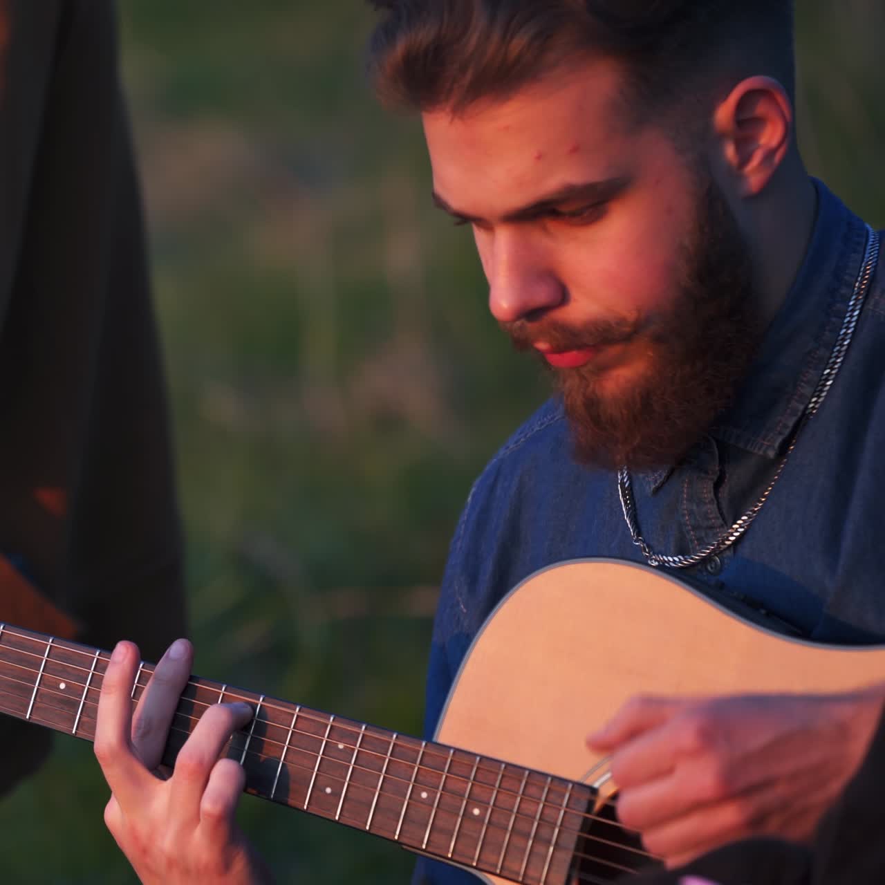 Young bearded man playing guitar while sitting on the ground. Male musician surrounded by friends at the blurred nature backdrop