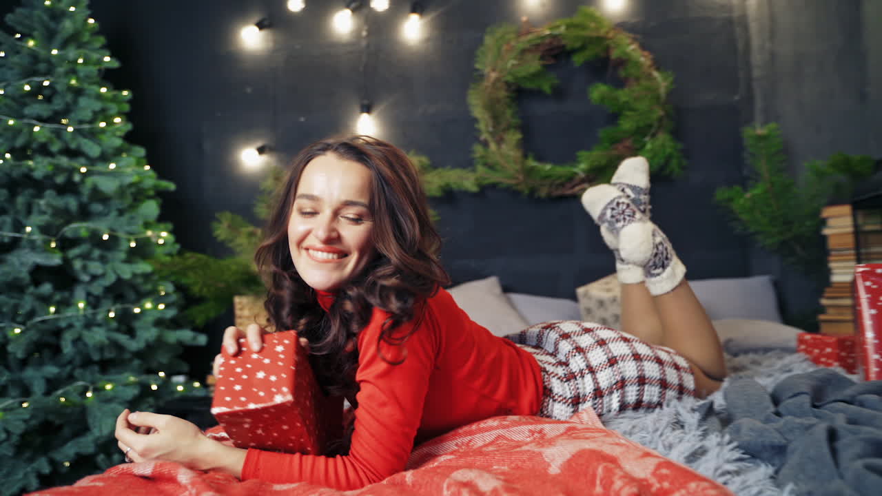 Woman in warm stylish clothes poses on bed in decorated room. Beauty in red sweater happy to receive gifts. Fest spirit.