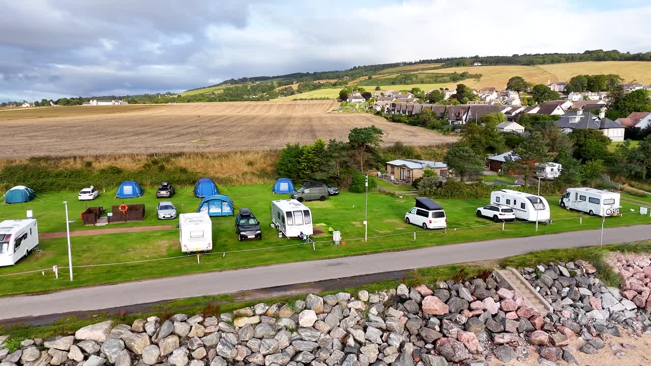 Drone footage glides over a coastal caravan and camping site in Rosemarkie, Scotland, showing motorhomes, tents, and cars on a bright, cloudy day
