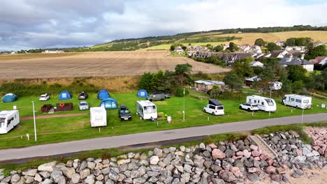drone footage glides over a coastal caravan and camping site in rosemarkie, scotland, showing motorhomes, tents, and cars on a bright, cloudy day