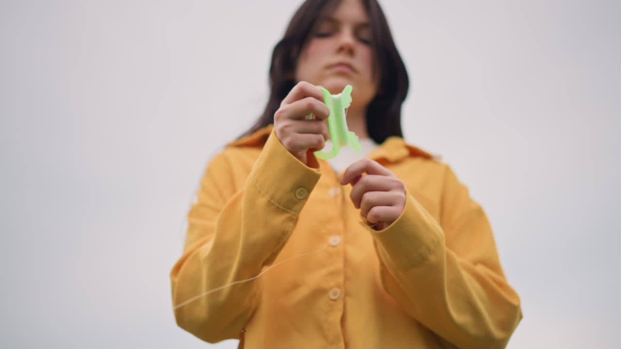 woman in yellow jacket stretching slime outdoors. Lowangle close shot of solitary figure pulling neon green slime between hands against overcast sky, textured jacket fabric, deliberate slow motions
