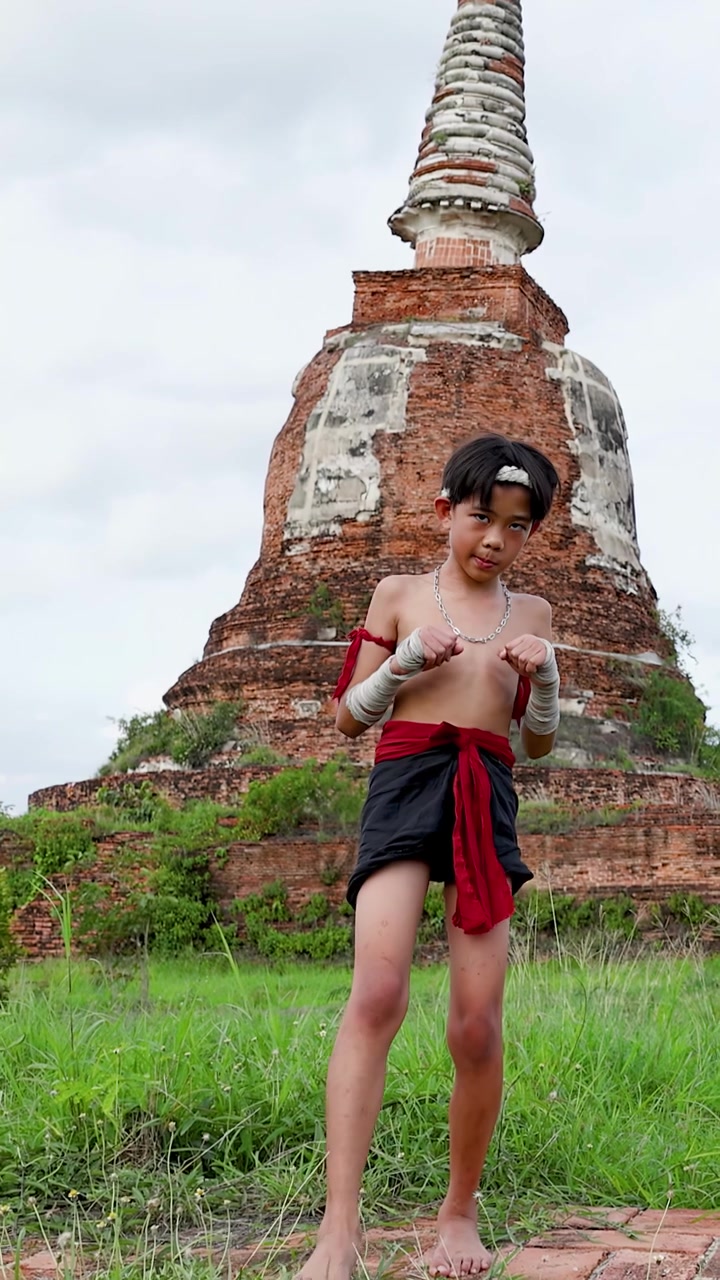 Menino Tailandês Executa Movimentos de Artes Marciais ao Ar Livre Perto de um Pagode Histórico Sob Céus Nublados
