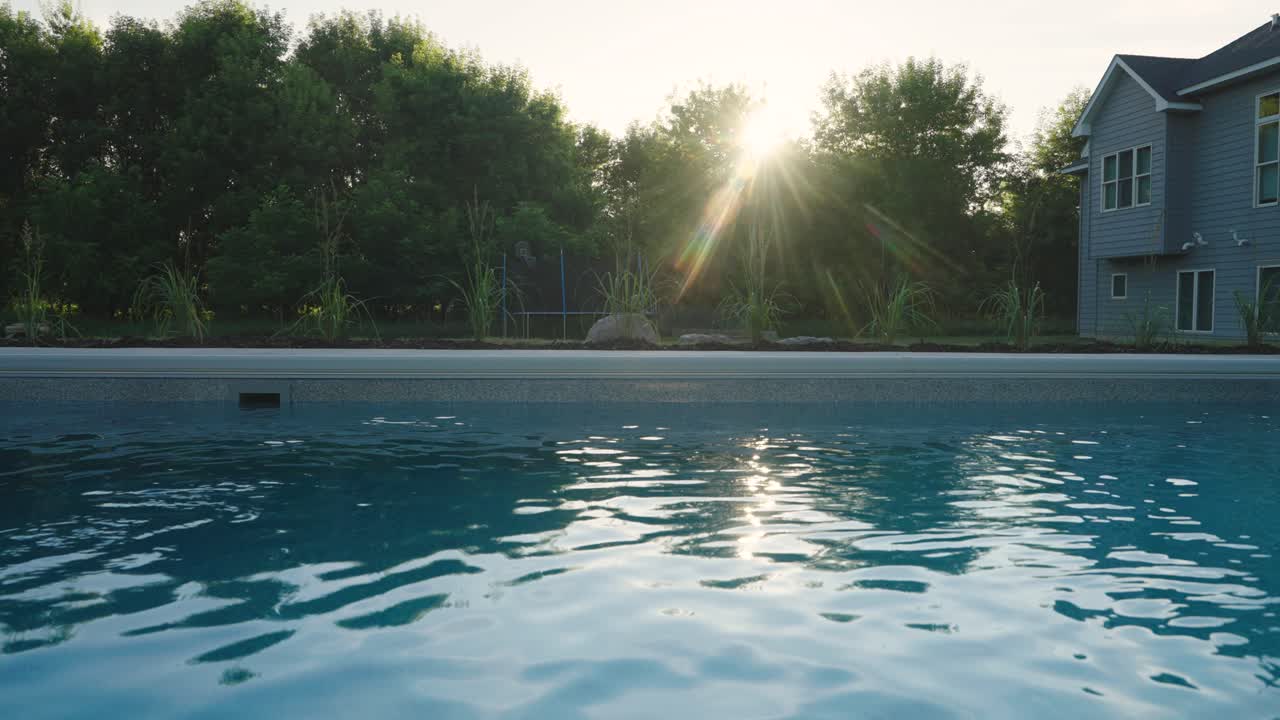Sunlight glimmers on fiberglass pool beside modern home with trees and lush landscaping