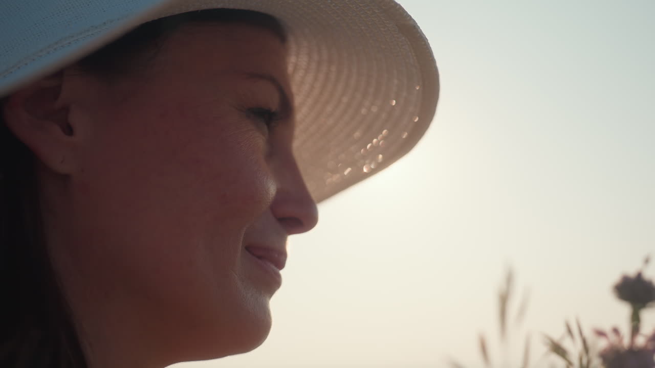 young woman in wide hat gently leans in to smell wildflowers in soft morning light, eyes closed in peaceful moment surrounded by natural bloom