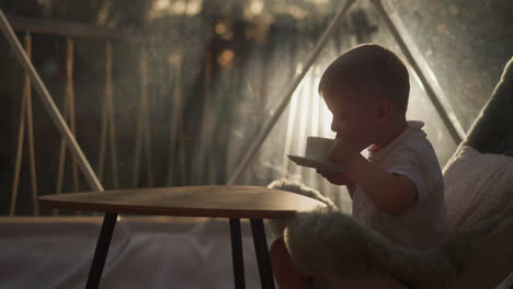 child takes sip of tea at low table at glamping. little boy drinks hot beverage sitting on floor in luxury camp. kid enjoys calmness on vacation