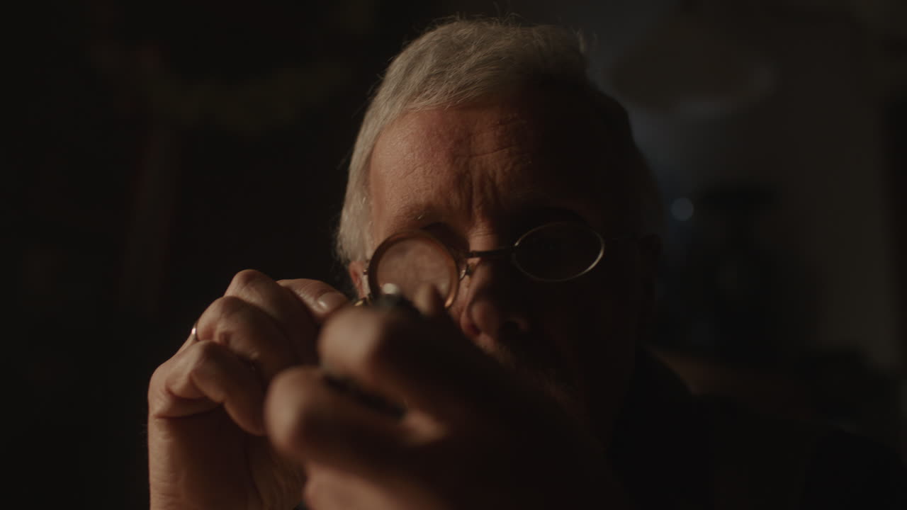 Older passionate man evaluates an old silver skull watch in his office at night