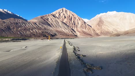 aerial drone shot of a car traveling beside a ladakhi river with snow-capped mountains in the distance.