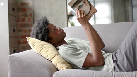 African-american-man-with-under-eye-patches-reading-book-on-sofa-at-home,-slow-motion