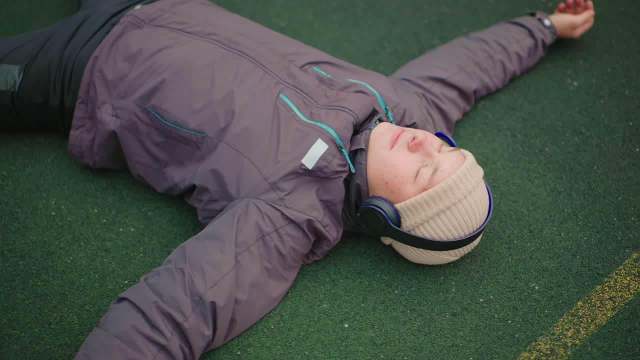 person lying on turf with headphones, wearing winter jacket and knitted beanie, eyes closed, arms spread, body relaxed after outdoor run, painted field line visible, ambient city hush, contemplative