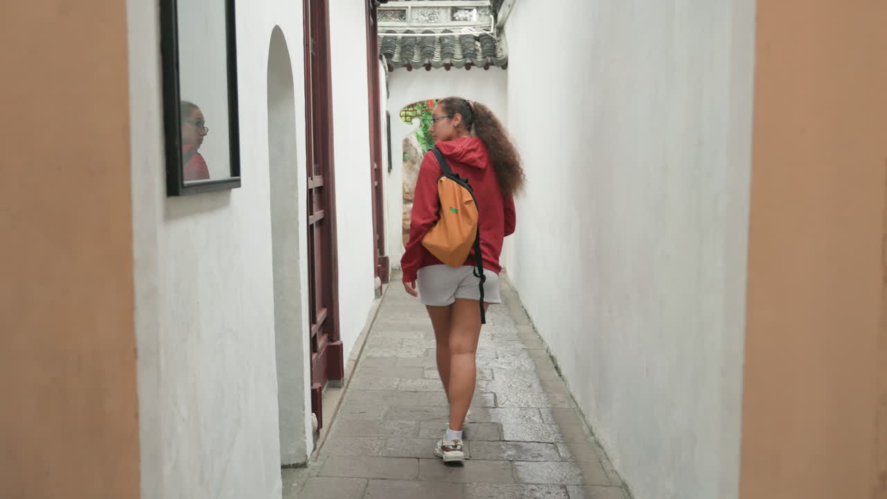 Giovane donna bianca che cammina in un vicolo stretto, maglione rosso e zaino giallo, pantaloncini di jeans e sneakers, che si allontana dalla macchina fotografica attraverso un arco e un sentiero piastrellato, tranquillo cortile storico, morbida naturale