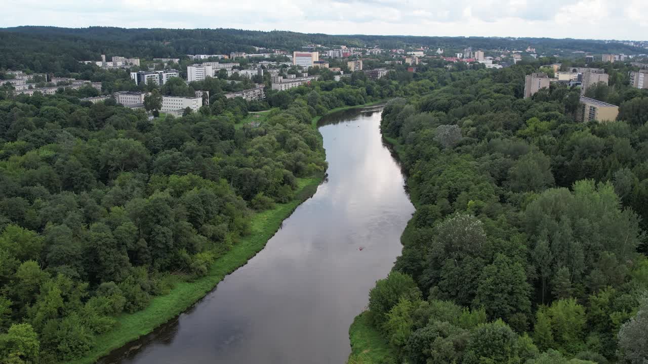 Drone shot of the Neris flowing through Vilnius, revealing dense summer trees along the bends and residential districts stretching deep into the Baltic capital