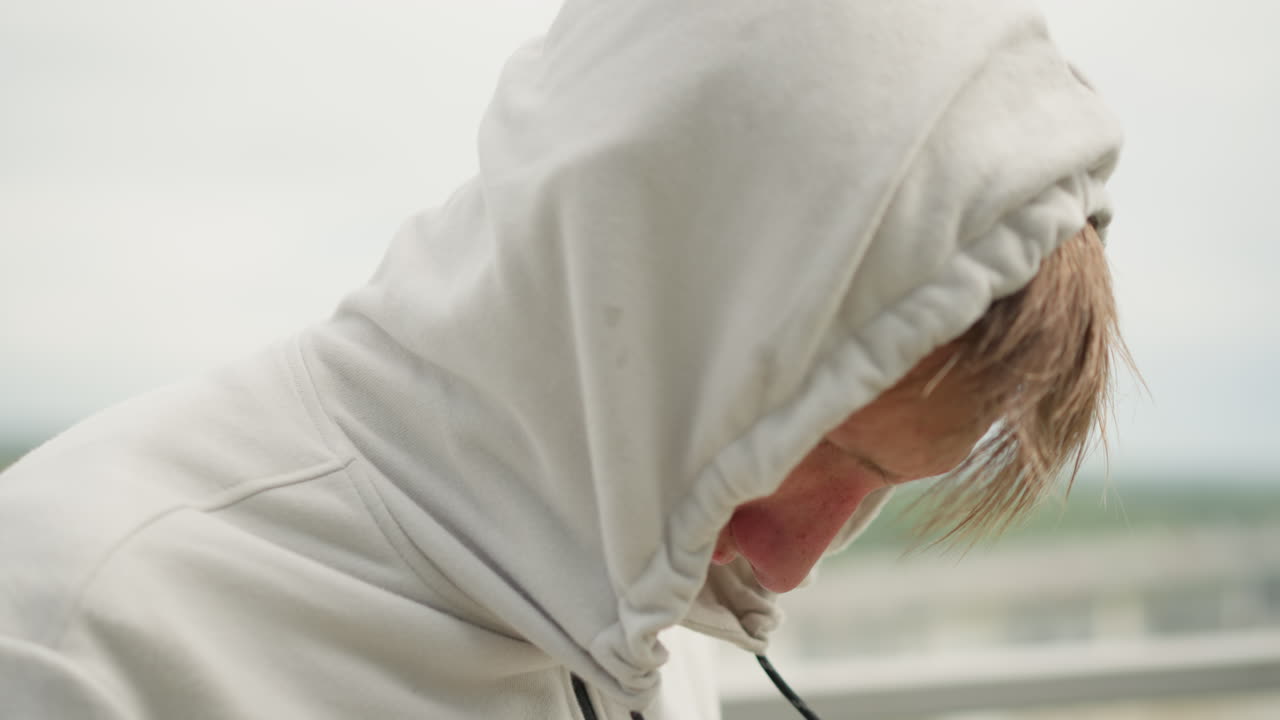 Man pulls hood tight, Overcast scene with concealed face, Closeup of man with hood over his face in gloomy weather, Man wearing hood stands on rooftop during overcast day with wet hair