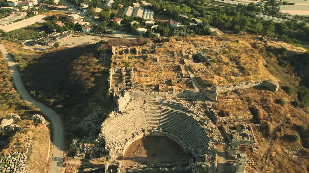 Drone video captures breathtaking aerial view of the ancient Greco-Roman theater ruins with tiered seating in hilly terrain near Patara, Turkey. Golden hues enhance historical aura