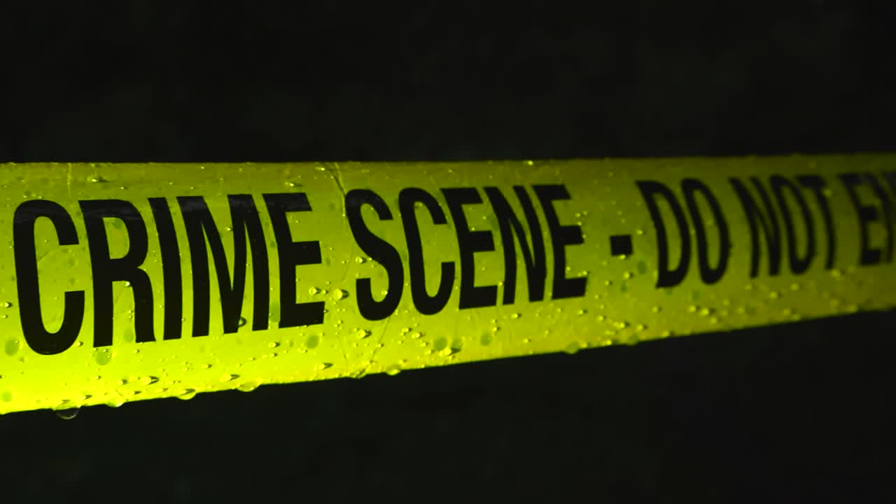 Police line yellow Crime Scene tape forensic investigation ribbon pulled infront of a dark night area while raindrops and water fall on it in slow motion at night. Spotlight light lighting the tape
