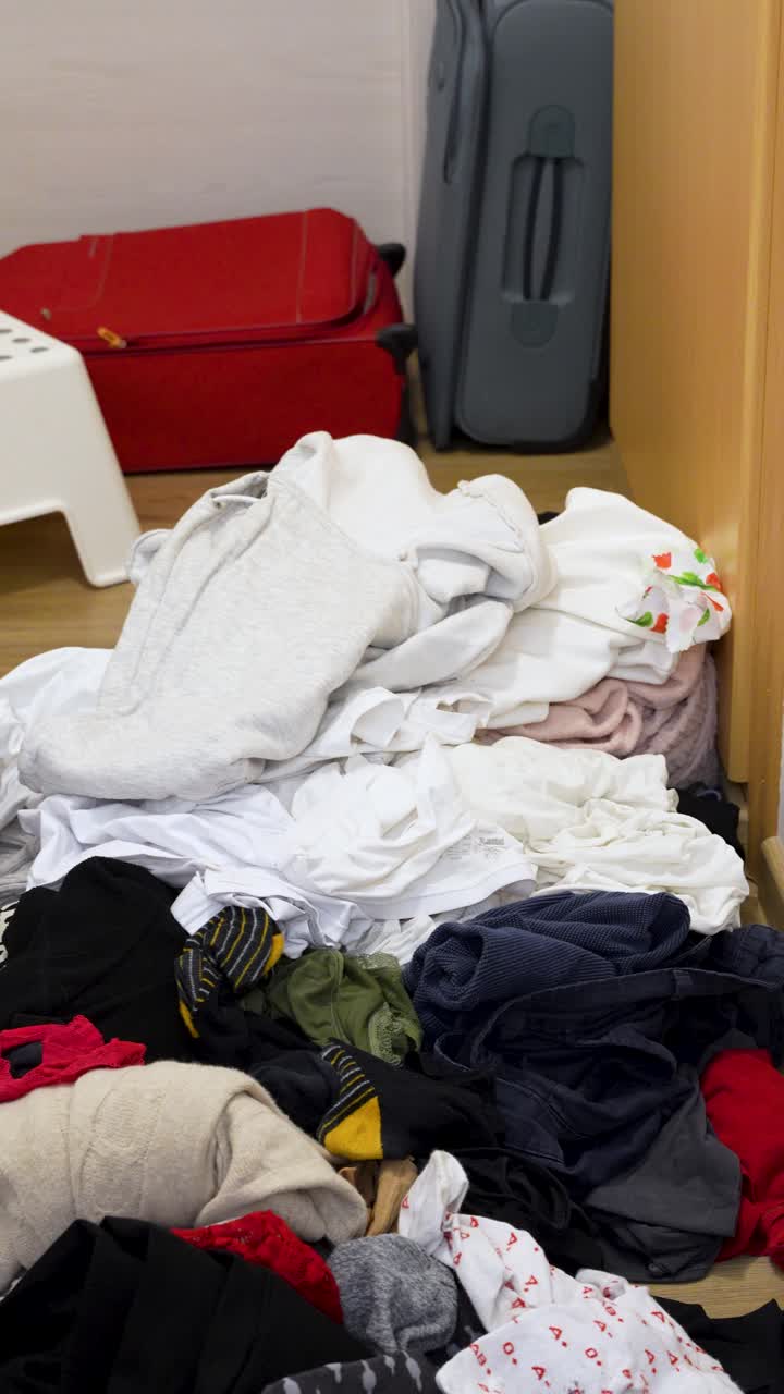 Messy pile of clothes on the floor with laundry baskets and an open suitcase nearby, showing post-travel laundry chaos and home cleanup after a trip. Vertical lifestyle scene