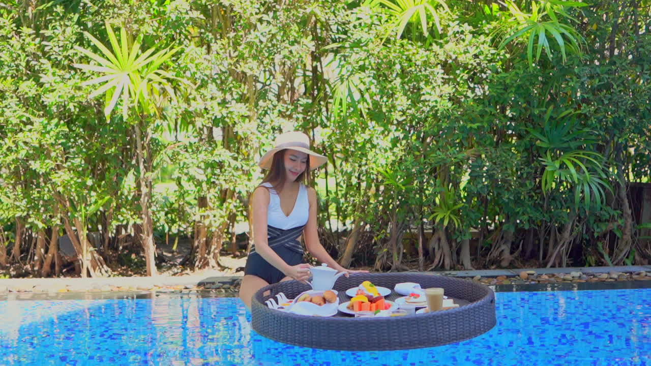 Slow-motion on sexy slim Asian model pouring hot tea in a cut. She is ready to eat tropical breakfast from floating on the water tray in a swimming pool