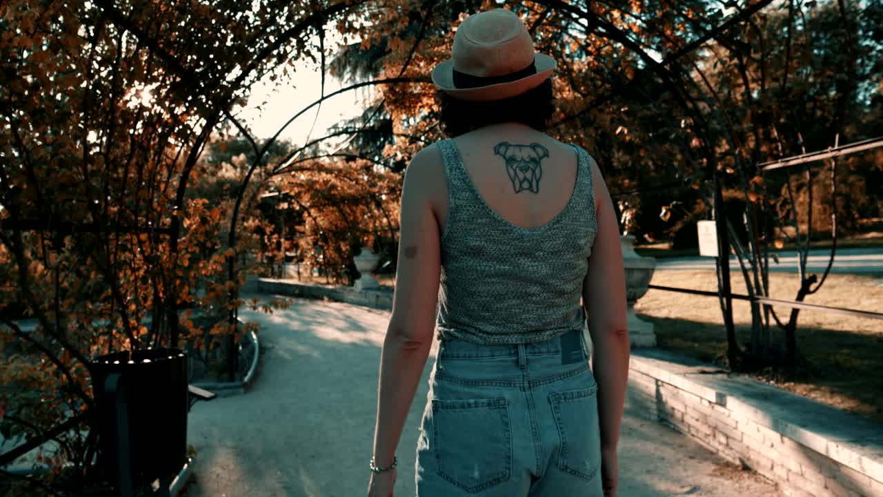 Mulher com Chapéu e Tatuagem Caminhando por uma Arcada de Parque Ensolarado